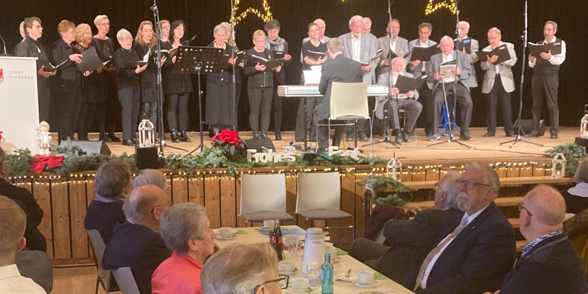 Weihnachtliche Feier in einer Halle. Auf der Bühne schwarz gekleidete Personen und davor sitzend Senioren