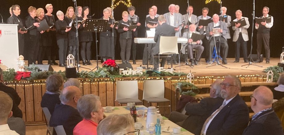 Weihnachtliche Feier in einer Halle. Auf der Bühne schwarz gekleidete Personen und davor sitzend Senioren