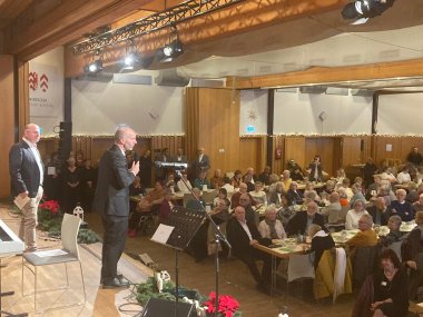Weihnachtliche Feier in einer Halle. Auf der Bühne ein Mann im Anzug mit einem Mikro in der Hand