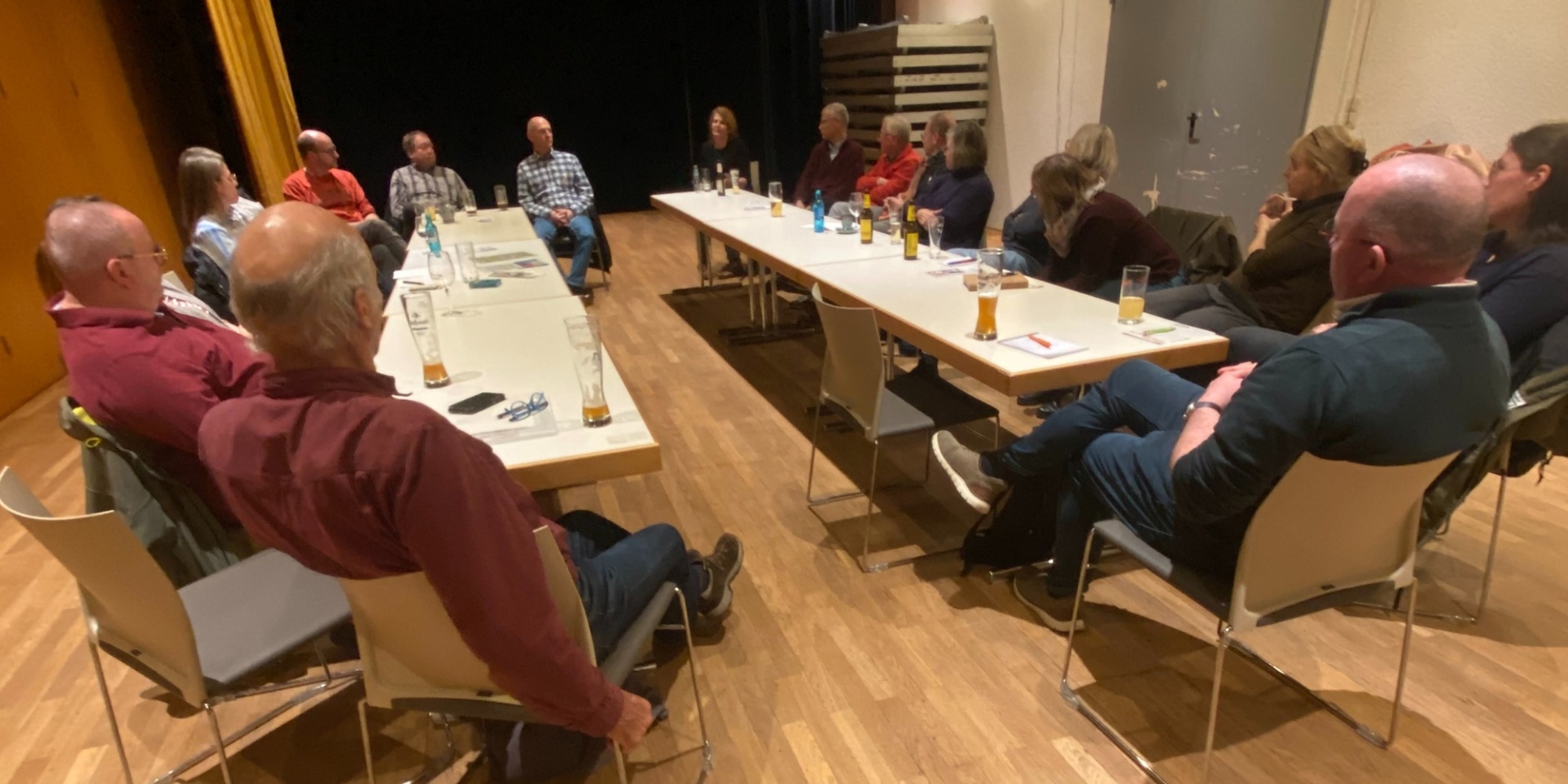 Mehrere Personen sitzen um zwei lange Tische. Vor einigen Personen sind Getränke im Glas abgestellt