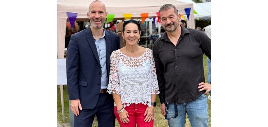 Eine Frau und zwei Männer stehend draußen. Im Hintergrund ein Zelt.