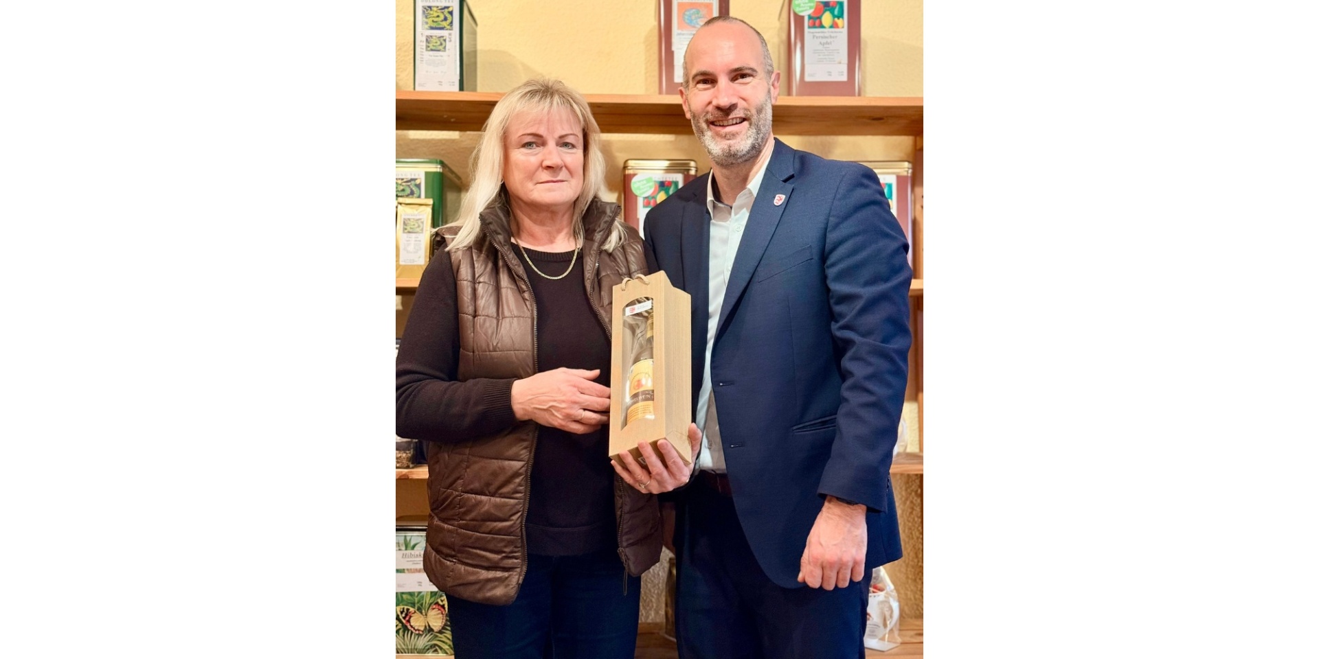 Ein Mann im Anzug steht neben einer blonden Frau. Die Frau hält eine verpackte Flasche als Geschenk in den Händen