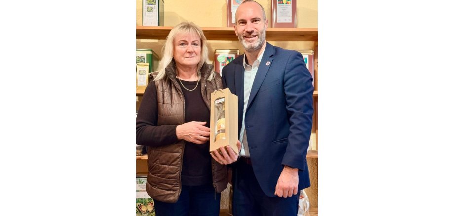 Ein Mann im Anzug steht neben einer blonden Frau. Die Frau hält eine verpackte Flasche als Geschenk in den Händen