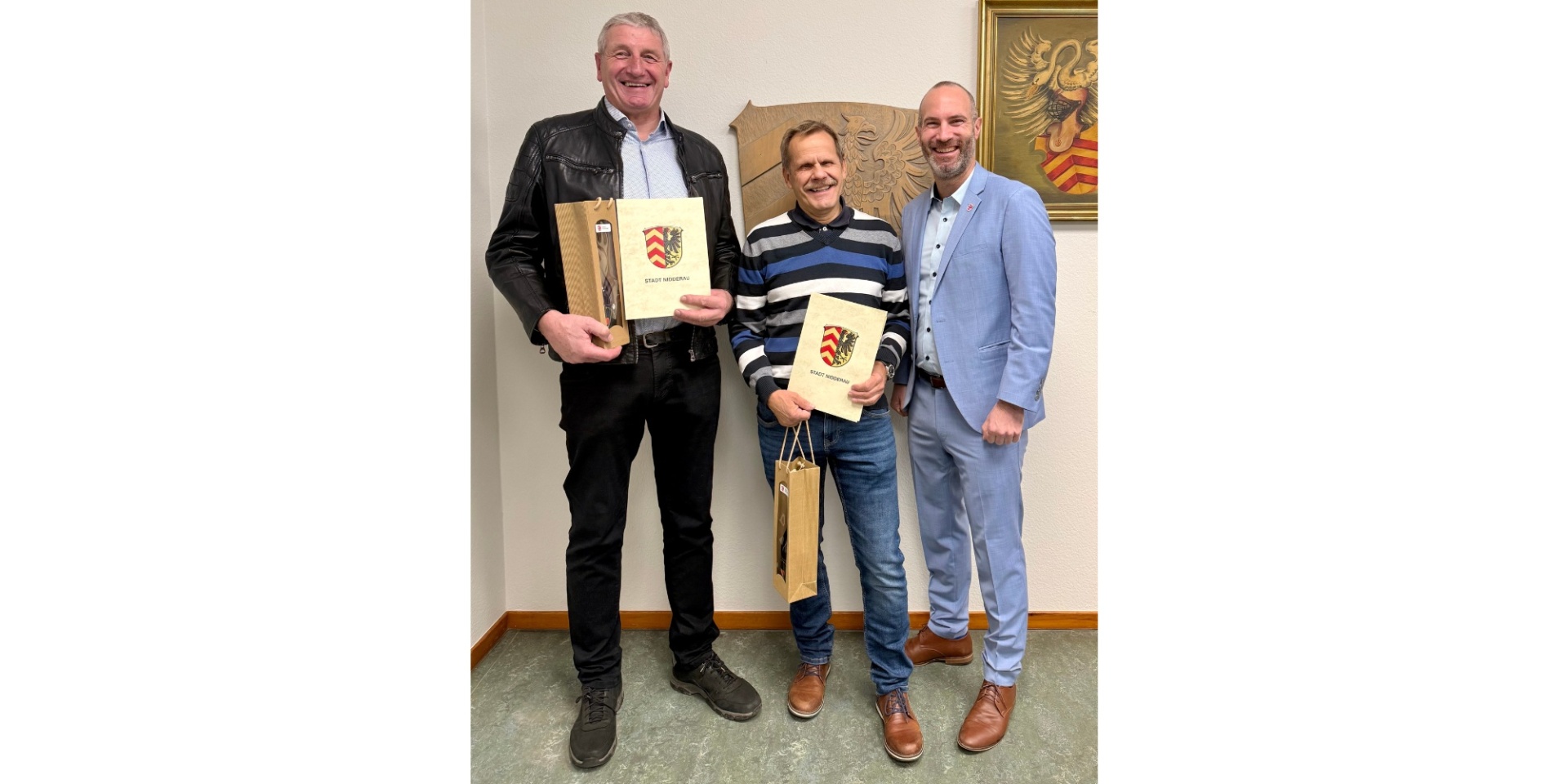 Drei Männer lächelnd und stehend nebeneinander. Zwei Männer halten je eine Urkunde und eine verpackte Flasche als Geschenk