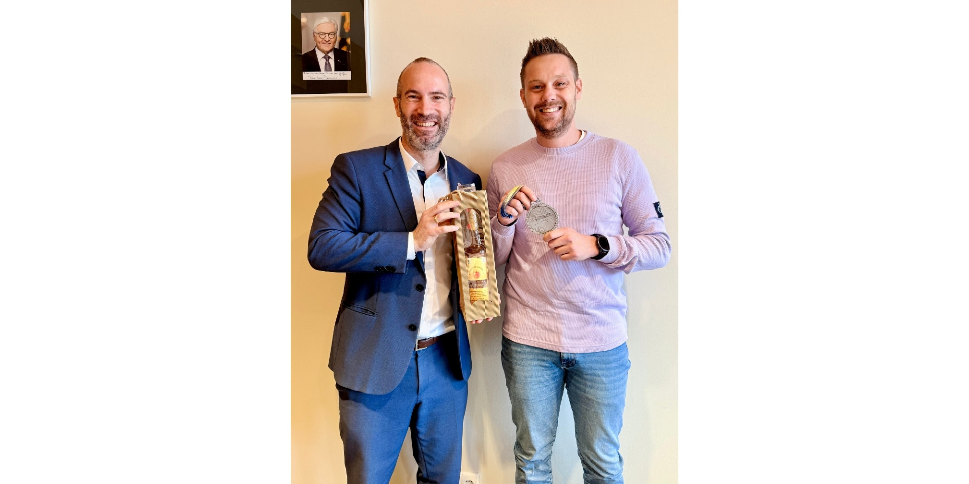 Zwei Männer stehend nebeneinander. Ein Mann hält eine Flasche und der andere eine Silbermedaille in den Händen