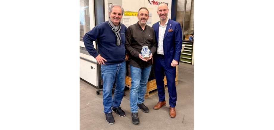 Drei Männer stehend. Der Mann in der Mitte hält einen Krug in den Händen.