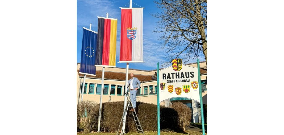 Ein Mann stehend auf einer Leiter. Im Hintergrund drei gehisste Flaggen und ein Gebäudekomplex.