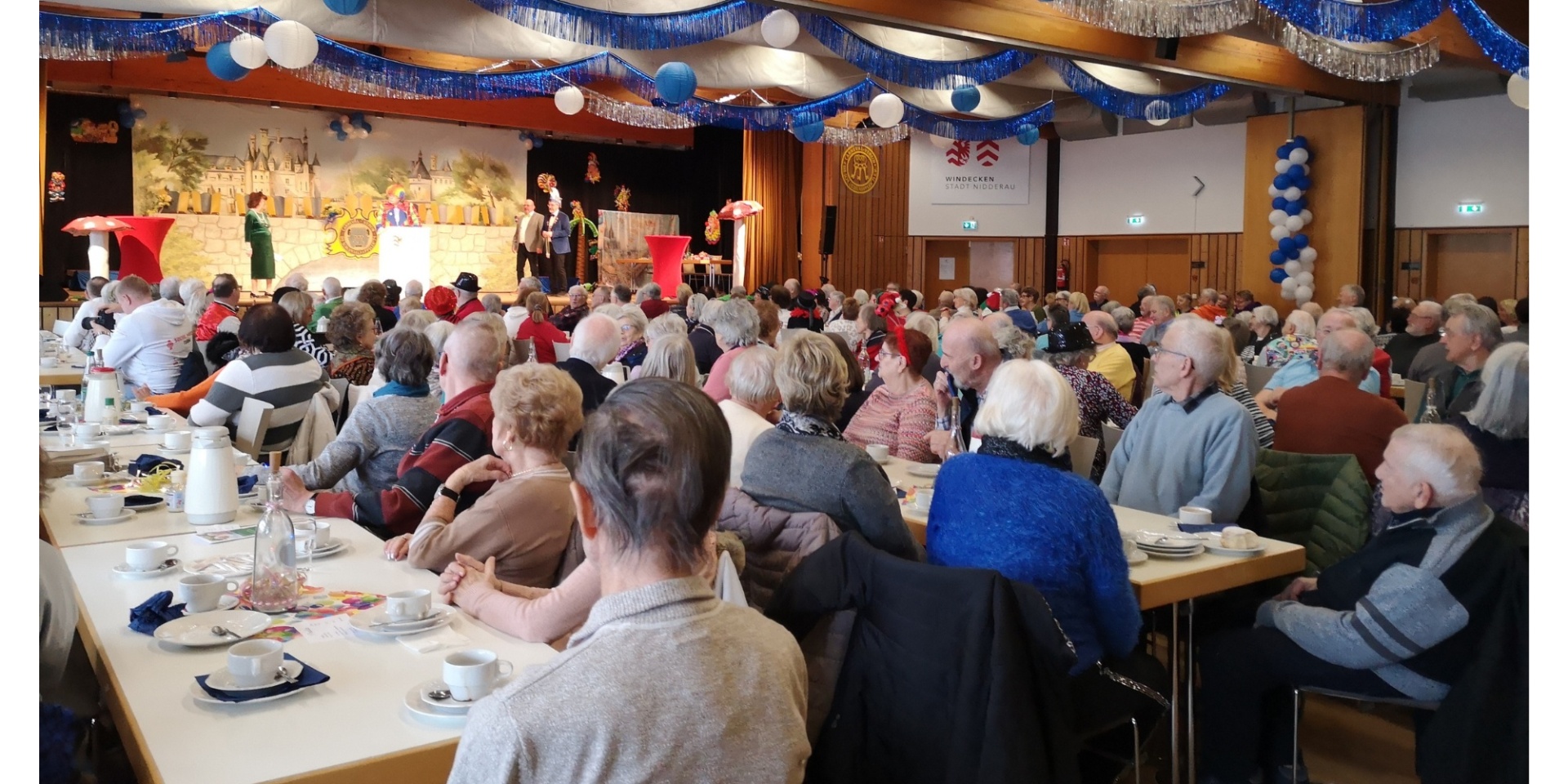 Gemischtes Publikum sitzend an Tischen in einem festlich geschmückten Veranstaltungssaal. Im Hintergrund eine Veranstaltung auf einer Bühne.