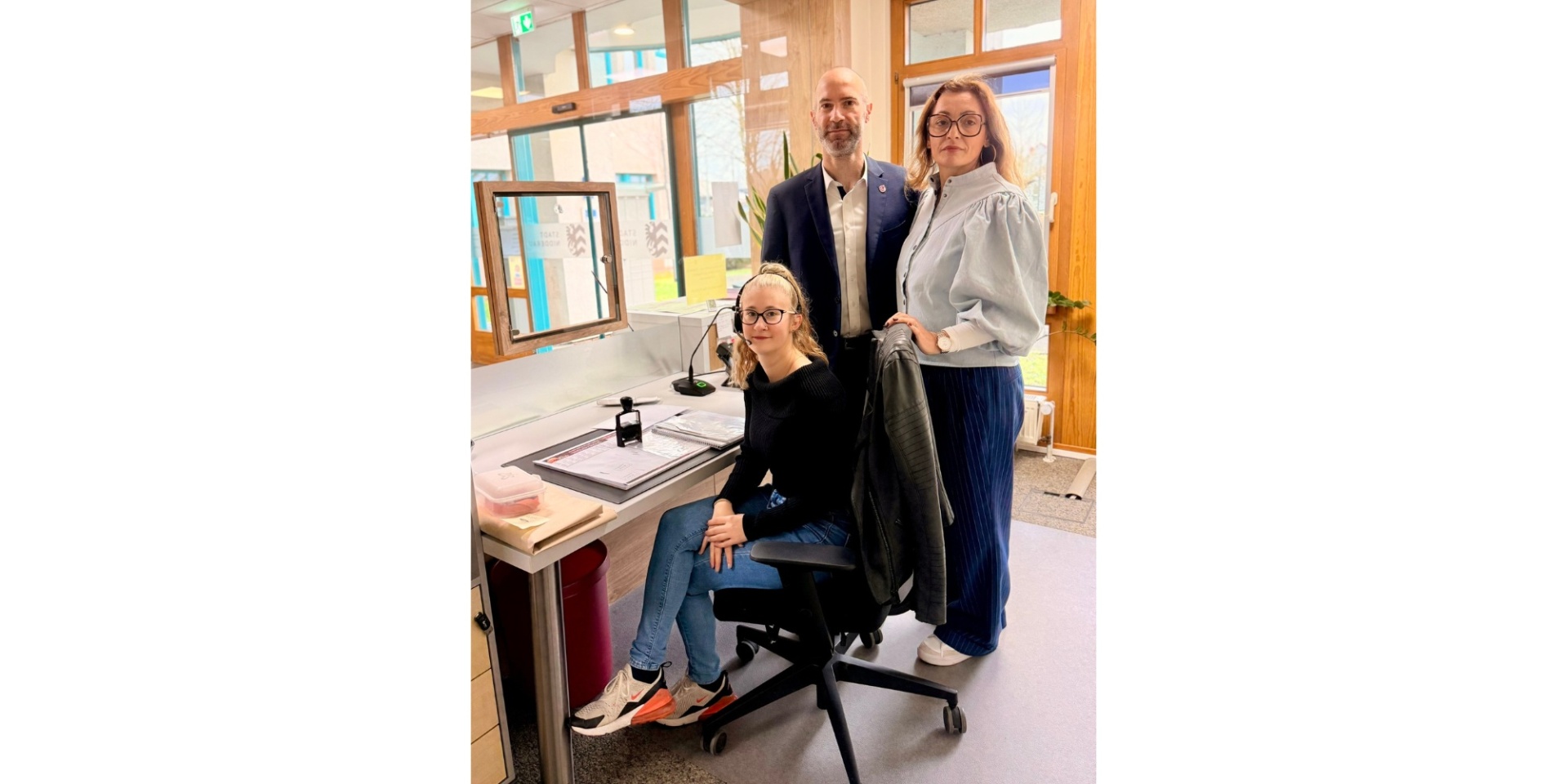 Eine Frau sitzend vor einem Tisch, dahinter ein Mann im Anzug und eine Frau stehend