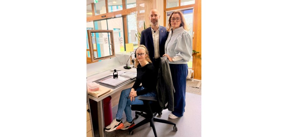 Eine Frau sitzend vor einem Tisch, dahinter ein Mann im Anzug und eine Frau stehend