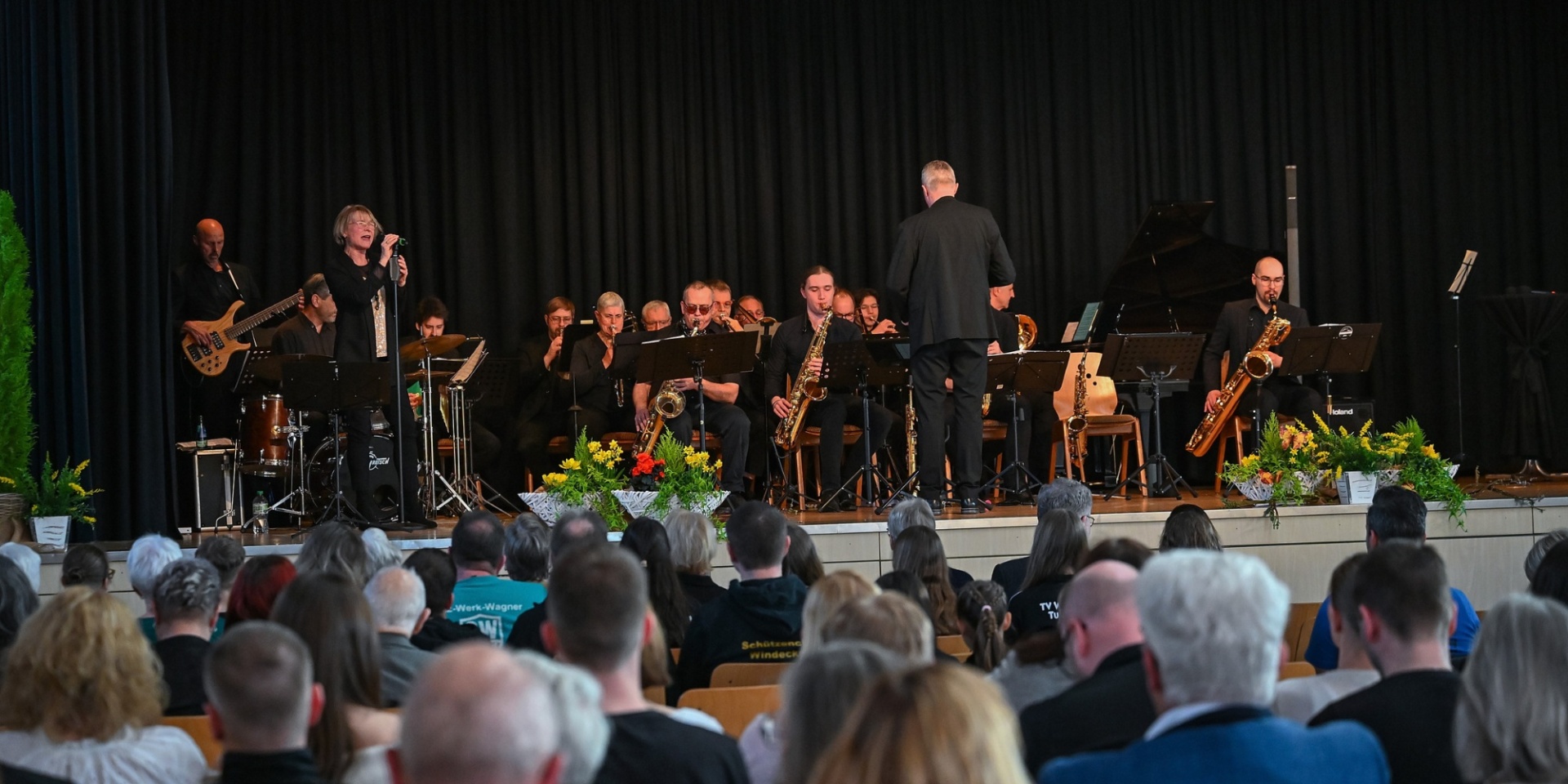 Schwarze Bühne mit Orchester davor sitzend ein Publikum