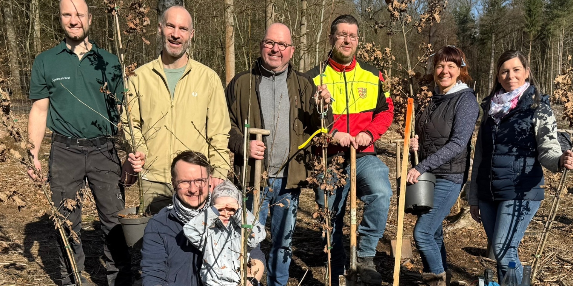 Mehrere Personen im Wald stehend oder kniend mit kleinen Baum-Setzlingen