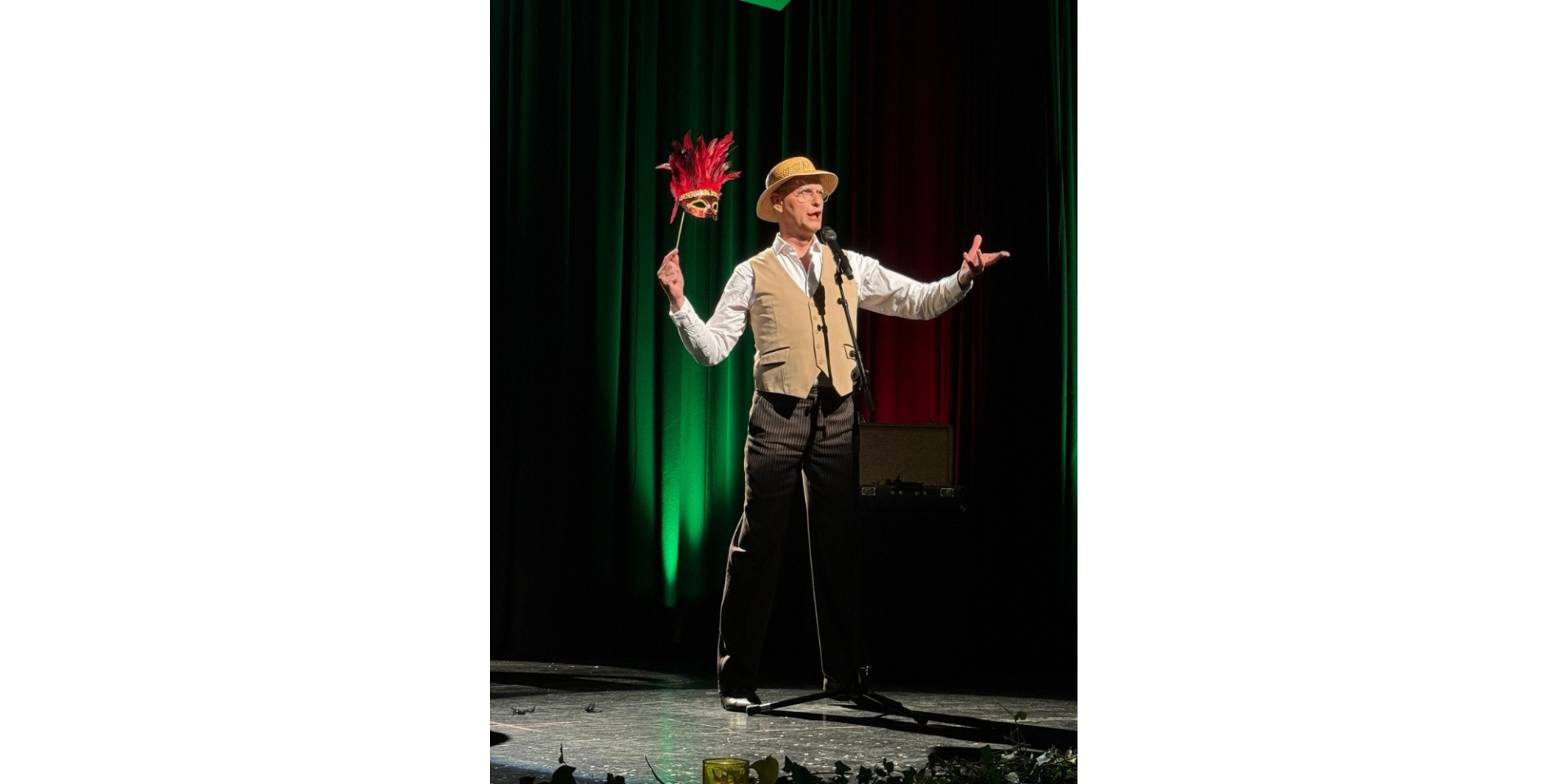 Mann auf einer schwarzen Bühne, in der einen Hand eine Maske haltend