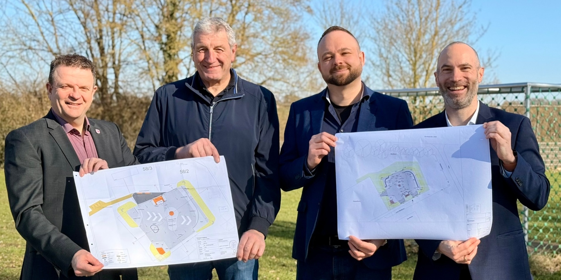 Vier Männer stehend auf einem Feld nebeneinander. Zwei davon halten jeweils einen Plan hoch