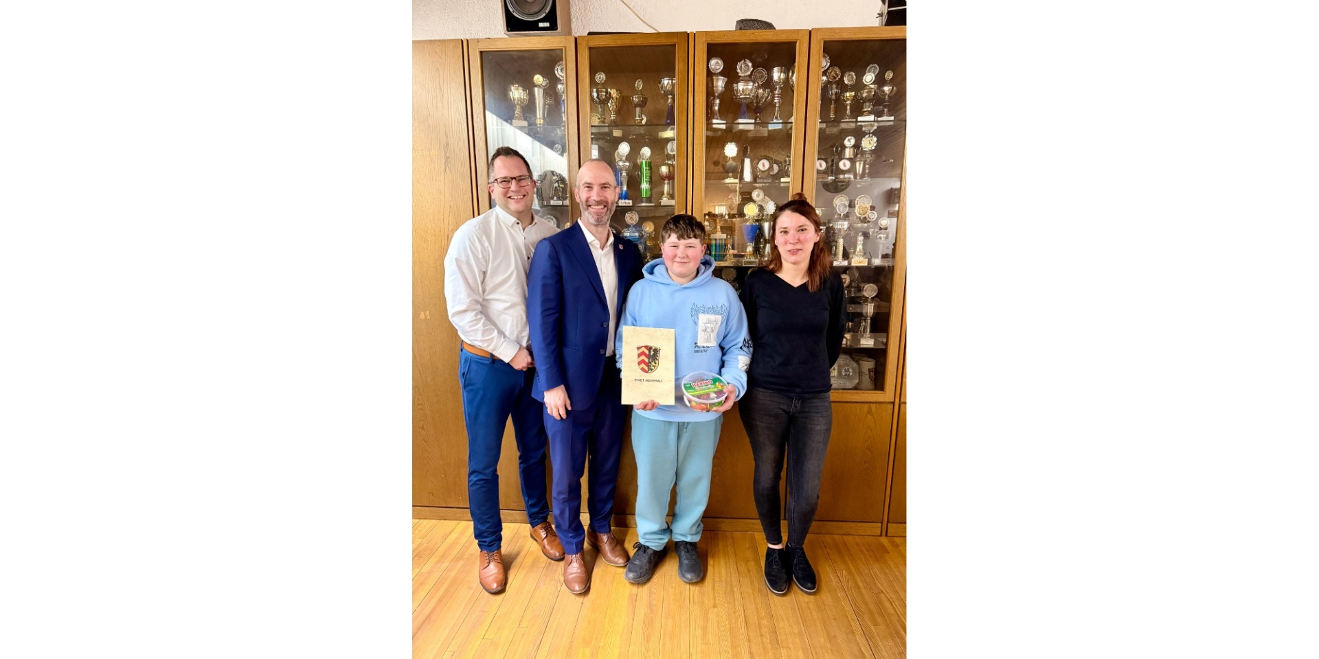 Vier Personen stehend in einem Raum. Ein Junge im Vordergrund hält eine Urkunde in der Hand