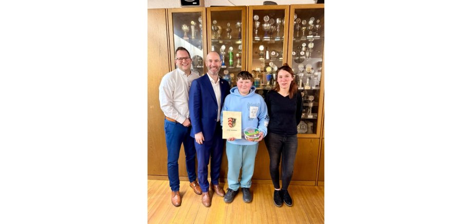 Vier Personen stehend in einem Raum. Ein Junge im Vordergrund hält eine Urkunde in der Hand