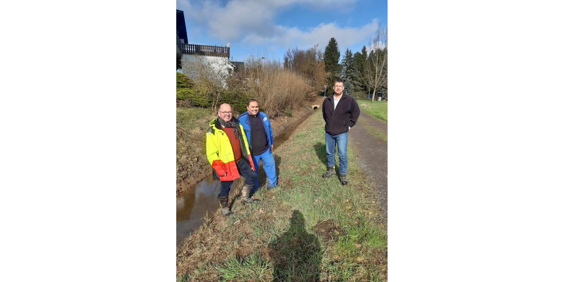Drei Personen stehend an einem Bachlauf auf einem Feld