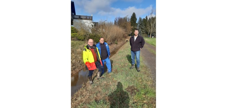 Drei Personen stehend an einem Bachlauf auf einem Feld