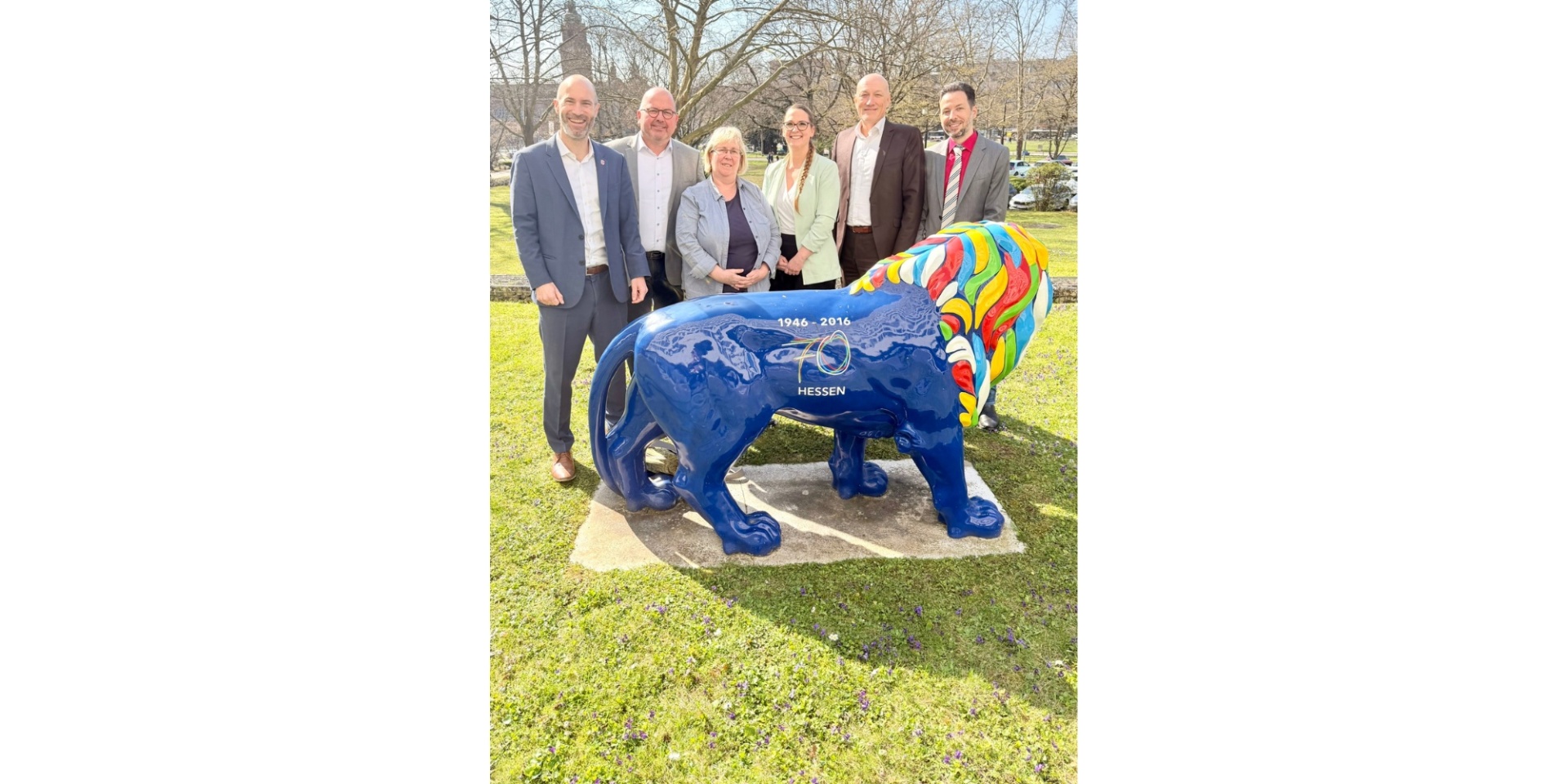 Vier Männer und zwei Frauen stehend auf einer Wiese hinter einer bunten Skulptur in Form eines Löwen