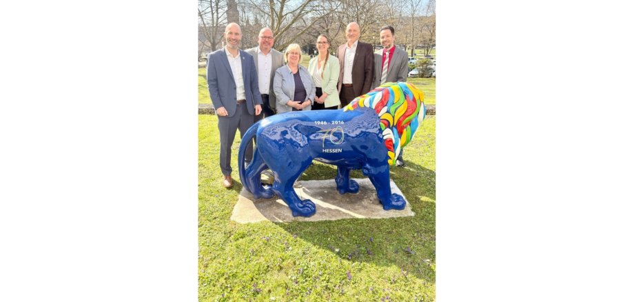 Vier Männer und zwei Frauen stehend auf einer Wiese hinter einer bunten Skulptur in Form eines Löwen