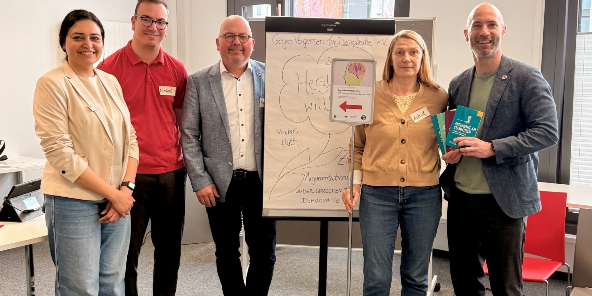 Fünf Personen stehend in einem Raum, in der Mitte ein Flipchart