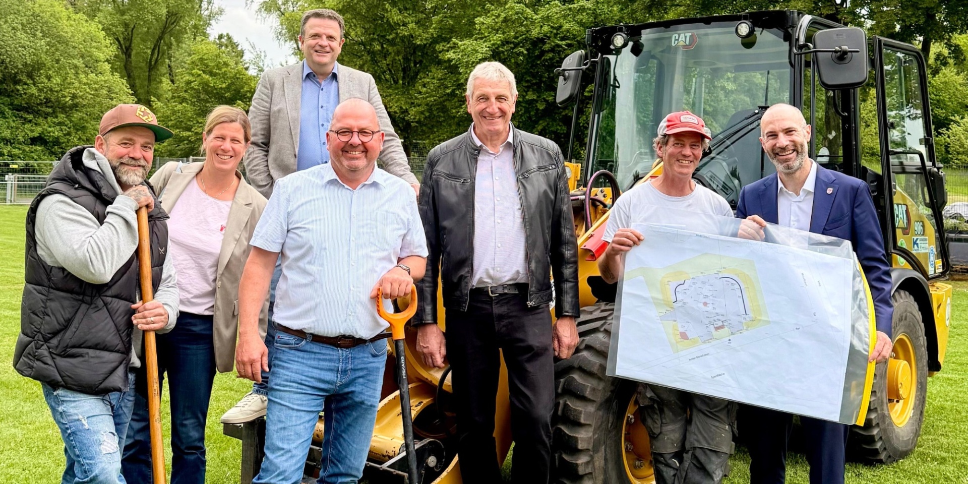 Mehrere Personen nebeneinander stehend auf einem Feld. Im Hintergrund steht ein Bagger