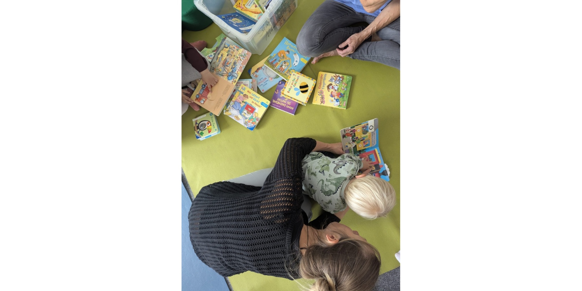 Frau beugt sich zu einem auf dem Boden sitzenden Kind hinunter. Auf dem Boden sind viele Kinderbücher verteilt