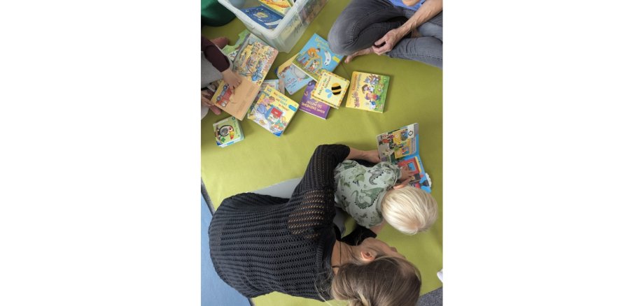 Frau beugt sich zu einem auf dem Boden sitzenden Kind hinunter. Auf dem Boden sind viele Kinderbücher verteilt