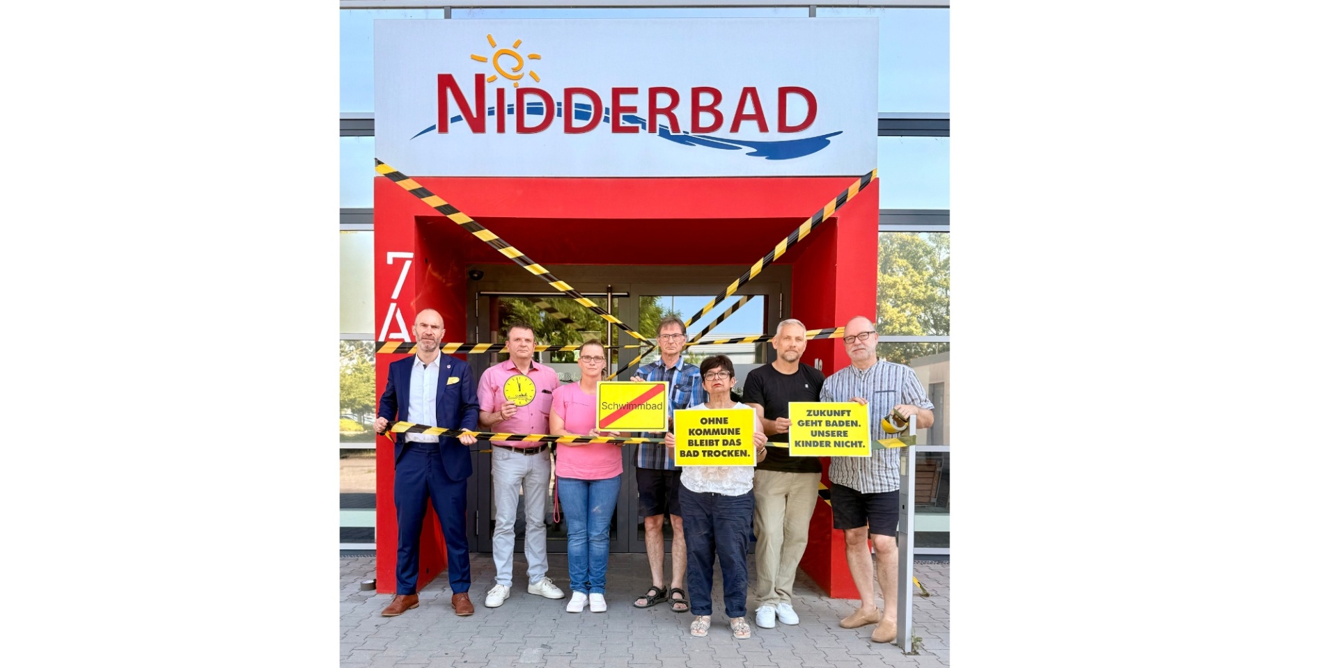 Mehrere Personen stehend vor dem Eingang eines Schwimmbads. Einige halten gelbe Schilder in der Hand
