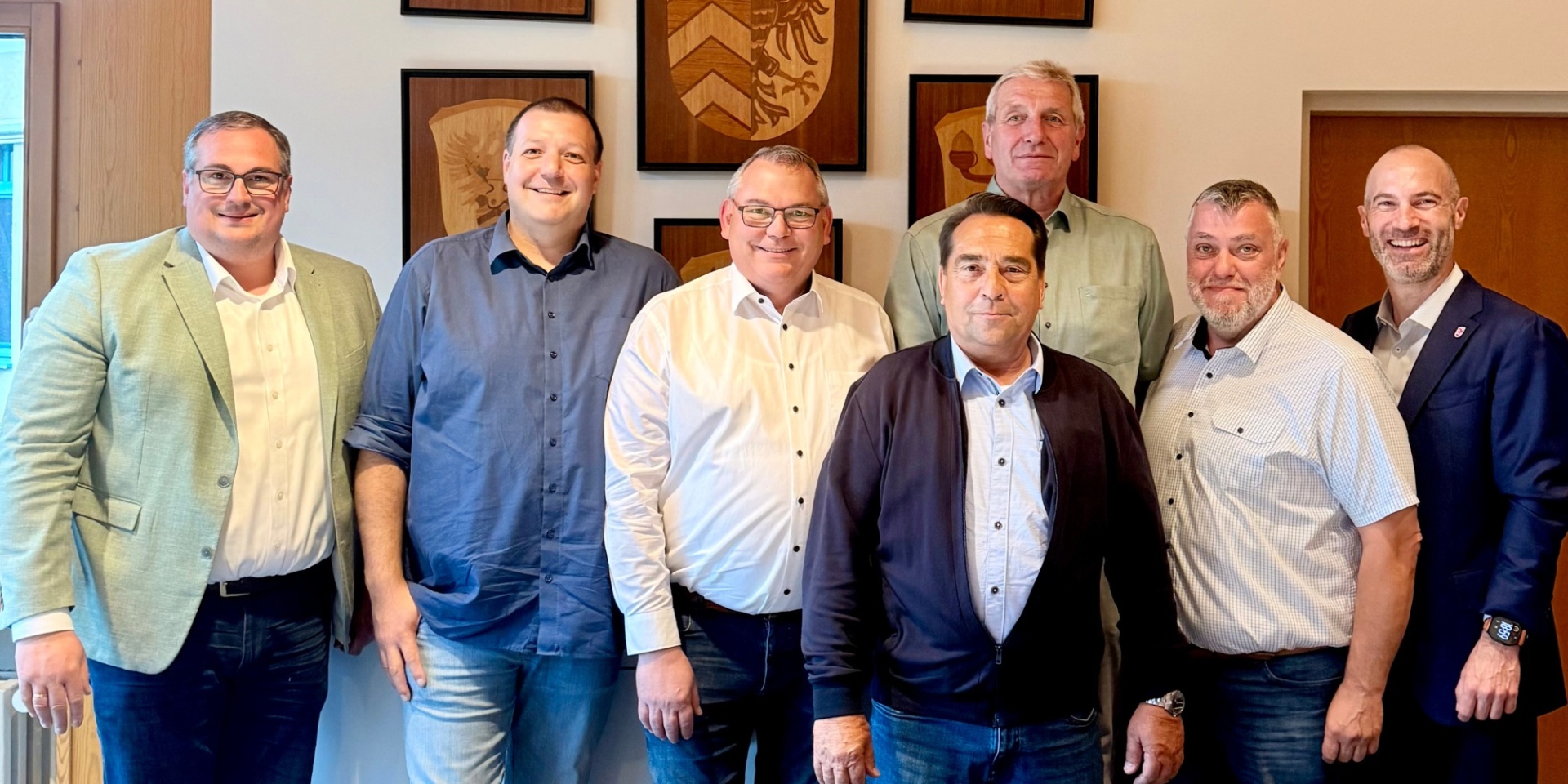 Sieben Männer stehend nebeneinander in einem Raum. Im Hintergrund sind Wappen aus Holz an der Wand angebracht