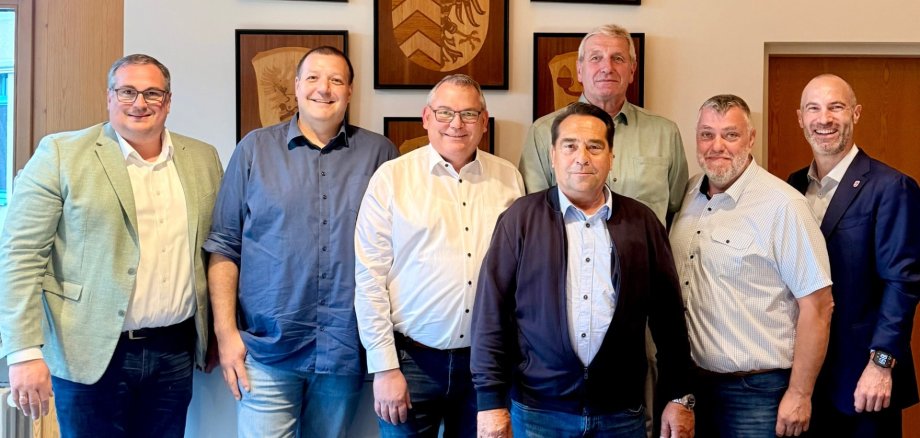 Sieben Männer stehend nebeneinander in einem Raum. Im Hintergrund sind Wappen aus Holz an der Wand angebracht