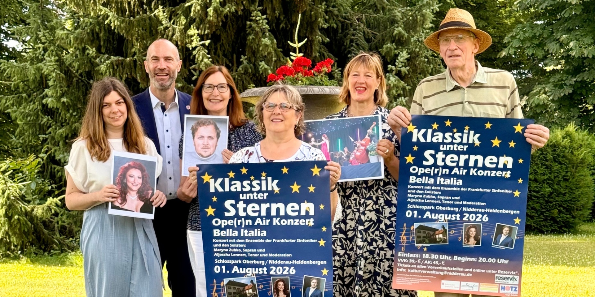 Vier Frauen und zwei Männer stehen nebeneinander in einem Park. Jeder hält entweder ein Veranstaltungsplakat oder ein Bild eines Künstlers in der Hand