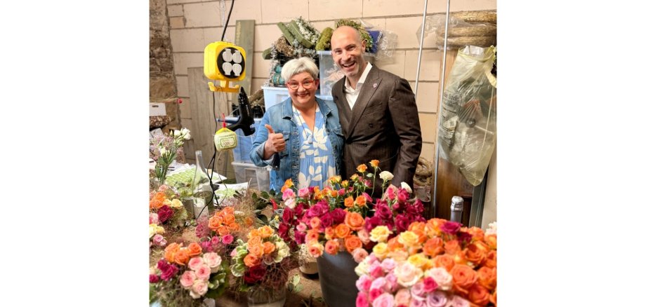 Eine Frau und eine Mann stehend. Im Vordergrund Blumensträuße.