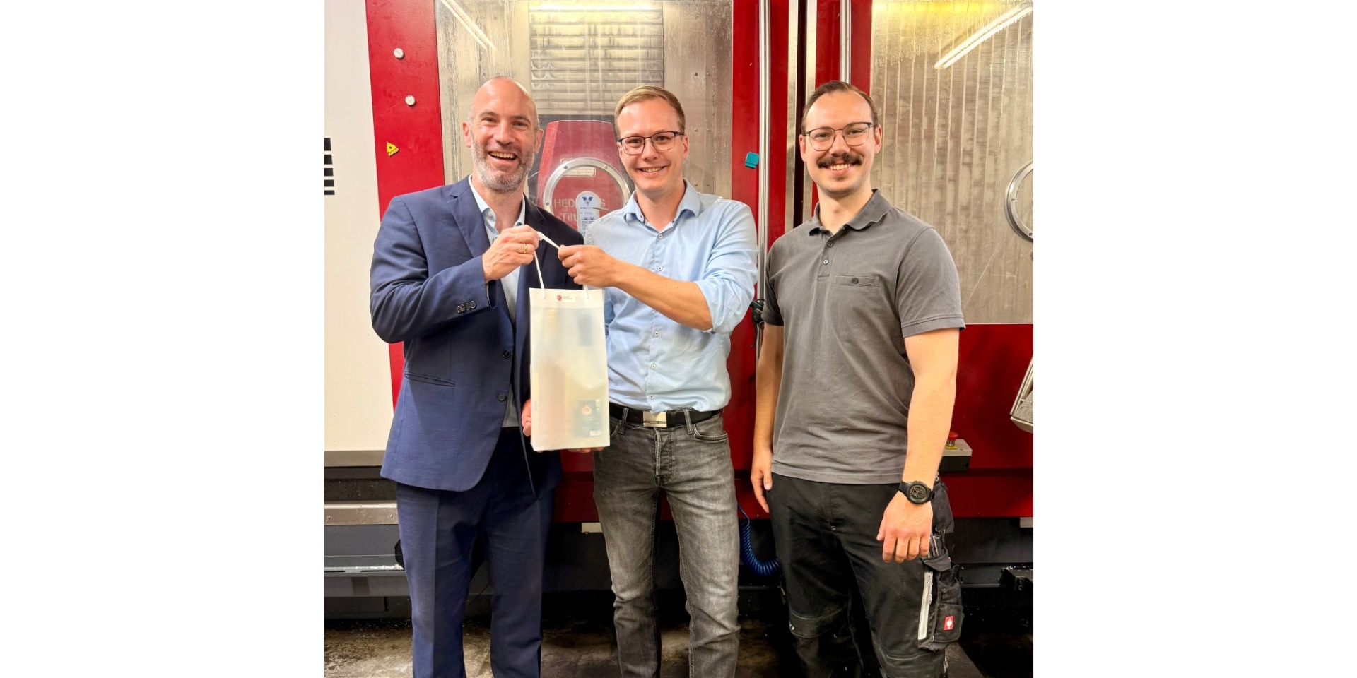 Drei Männer stehend. Ein Mann hält eine Präsenttasche in der Hand.