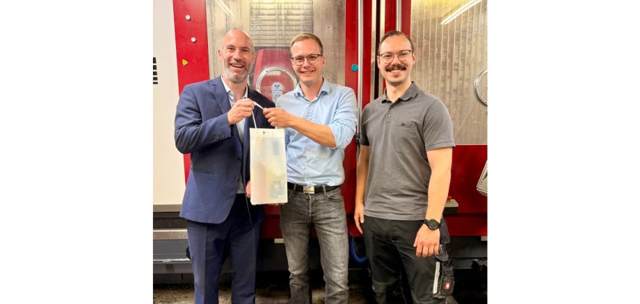 Drei Männer stehend. Ein Mann hält eine Präsenttasche in der Hand.