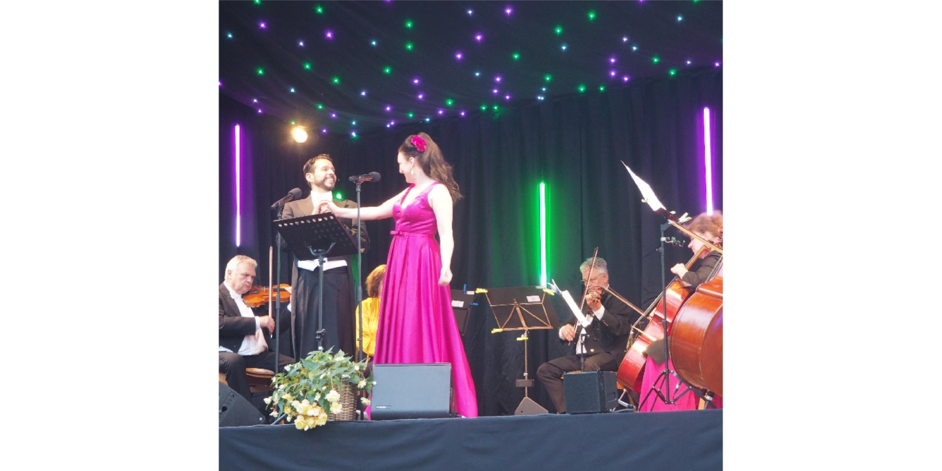 Eine Frau und ein Mann auf einer Bühne. Ein Orchester spielt im Hintergrund Instrumente.