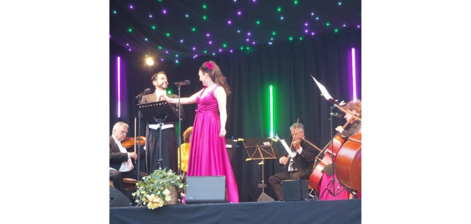Eine Frau und ein Mann auf einer Bühne. Ein  Orchester spielt im Hintergrund Instrumente.