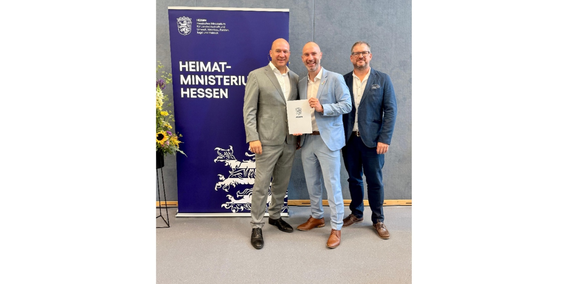 Drei Männer stehend vor einem großen Aufstellplakat.