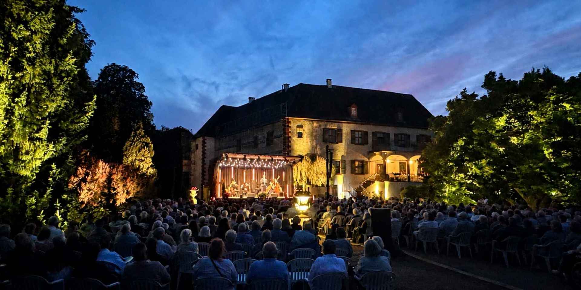 In einem Schlosspark sitzen viele Gäste auf Stühlen im Freien und schauen auf eine Bühne vor einer Schlossfassade
