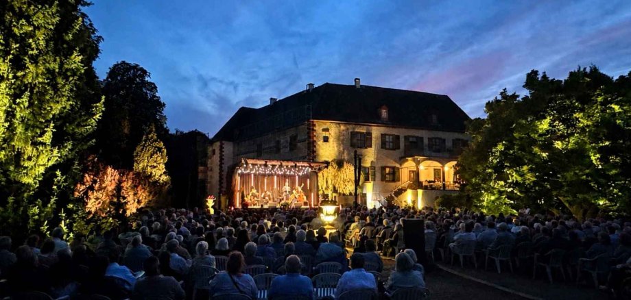 In einem Schlosspark sitzen viele Gäste auf Stühlen im Freien und schauen auf eine Bühne vor einer Schlossfassade