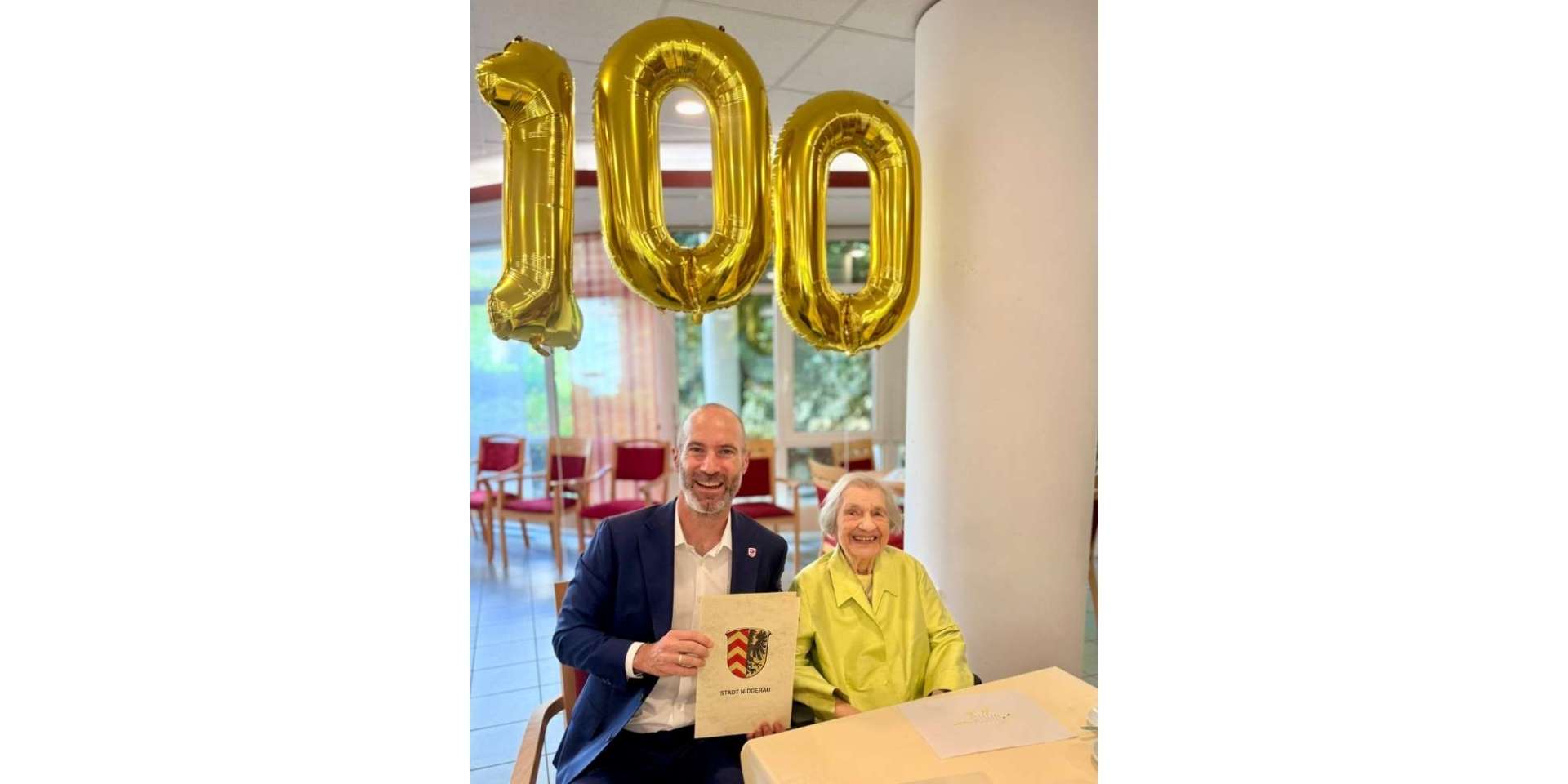 Ein Mann im Anzug sitzend neben einer älteren Frau. Im Hintergrund Luftballons zum 100. Geburtstag