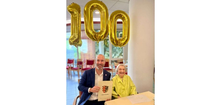 Ein Mann im Anzug sitzend neben einer älteren Frau. Im Hintergrund Luftballons zum 100. Geburtstag