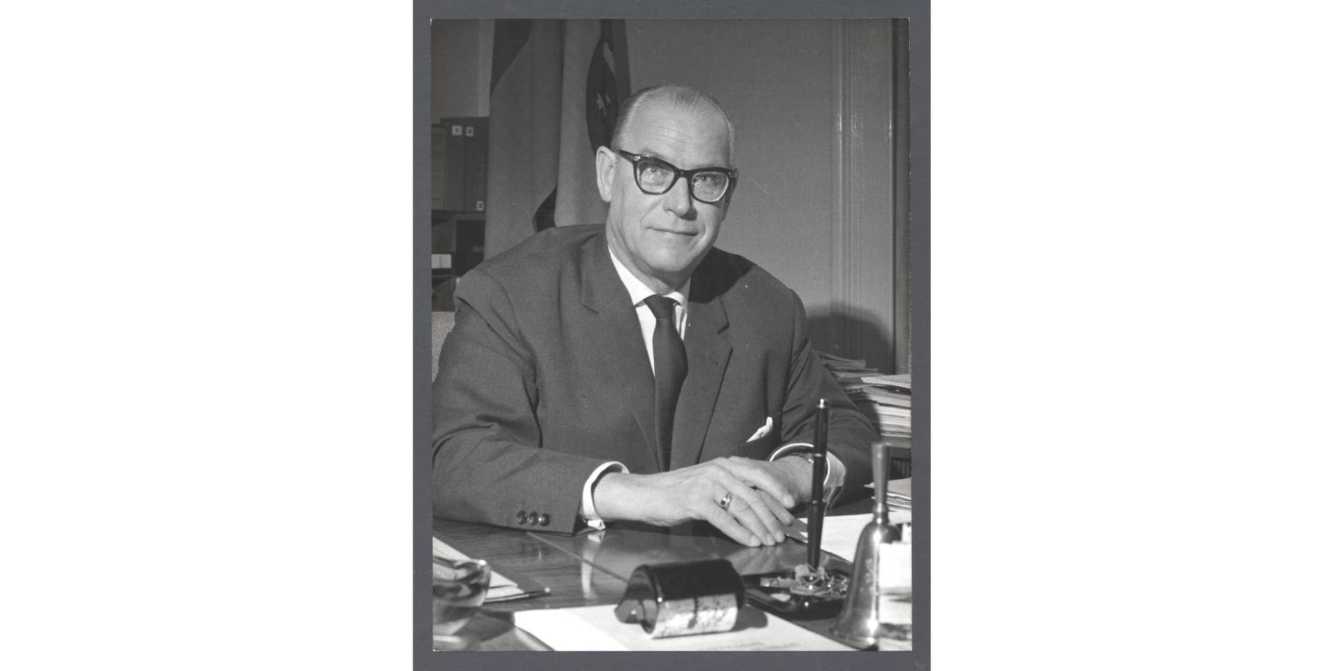 Schwarz-weiß Fotografie eines Mannes mit Brille sitzend im Anzug an einem Schreibtisch