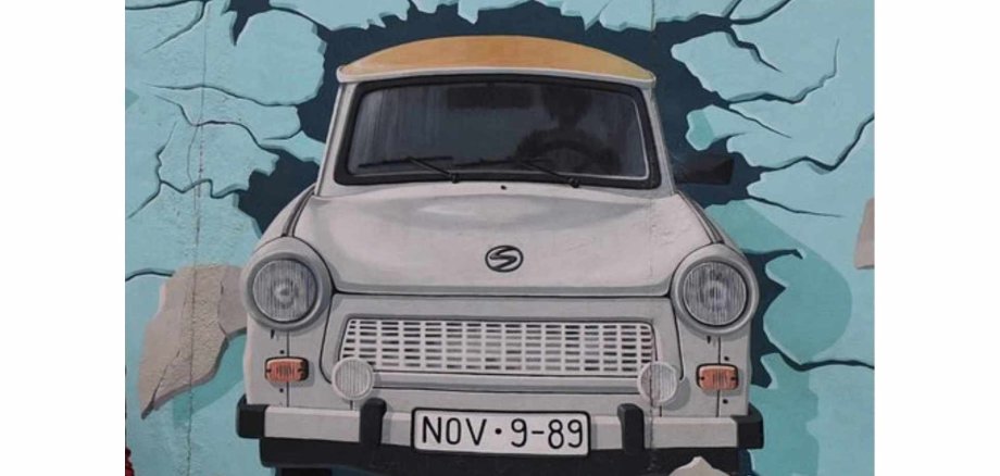 Bild an der Berliner Mauer: Grauer Trabbi durchbricht eine blaue Wand