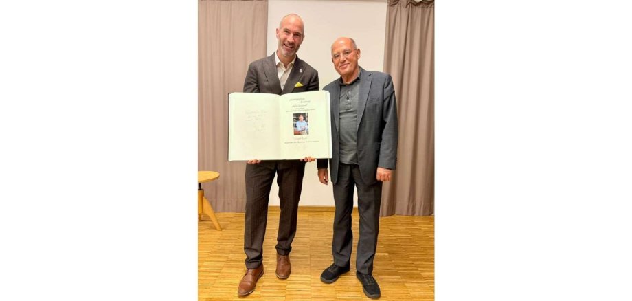 Zwei Männer in Anzügen stehend nebeneinander. Ein Mann hält ein geöffnetes Buch vor sich