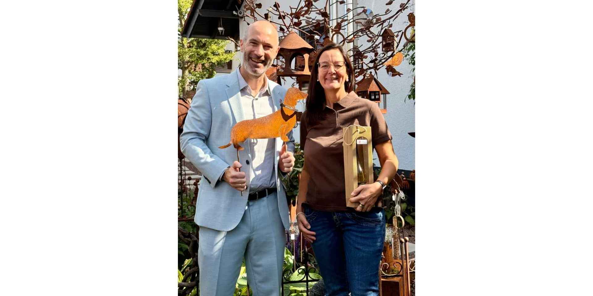 Ein Mann und eine Frau stehen in einem Garten. Der Mann hält eine Dackelskulptur aus Stahl