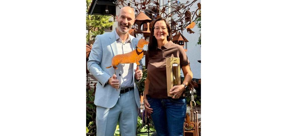 Ein Mann und eine Frau stehen in einem Garten. Der Mann hält eine Dackelskulptur aus Stahl
