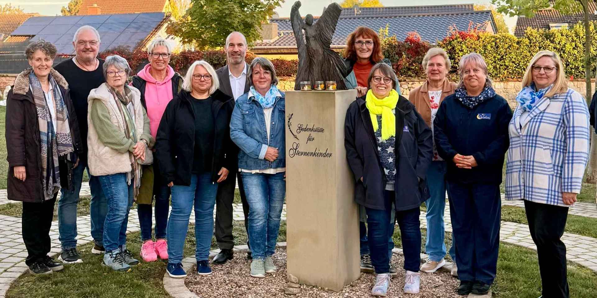 Mehrere Personen stehen nebeneinander auf einem Friedhof. In der Mitte befindet sich ein Gedenkstein für Sternenkinder