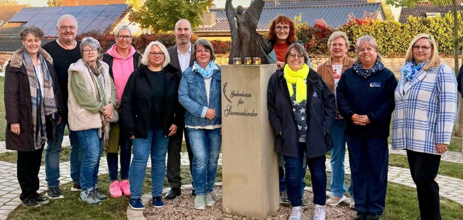 Mehrere Personen stehen nebeneinander auf einem Friedhof. In der Mitte befindet sich ein Gedenkstein für Sternenkinder
