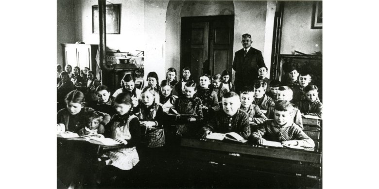 Schwarz-weiß Aufnahme aus einem Klassenraum mit jeweils zwei Schülern sitzend auf einer Bank. Im Hintergrund steht der Lehrer im Anzug